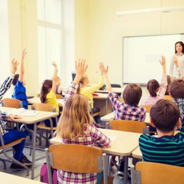 Em uma sala de aula, alunos sentados em suas cadeiras, levantam a mão.