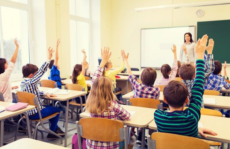 Em uma sala de aula, alunos sentados em suas cadeiras, levantam a mão.