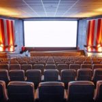 Foto de uma sala de cinema com a tela branca e poltronas vazias.