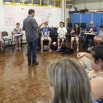 Oficina - Em uma sala aula, alunos em semi-círculo sentados em cadeiras de frente para o professor Luiz Amorim. Professor está em pé de costas para a foto e de frente para os alunos.