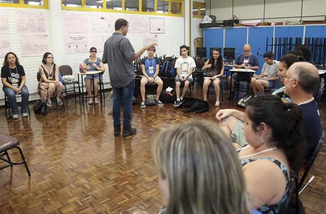 Oficina - Em uma sala aula, alunos em semi-círculo sentados em cadeiras de frente para o professor Luiz Amorim. Professor está em pé de costas para a foto e de frente para os alunos.