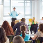 Fotografia colorida de uma sala de aula repleta de alunos e o professor. Os alunos, em primeiro plano, estão sentados de costas para a foto e de frente para o professor. O professor, em imagem desfocada, está de frente para os alunos com os braços flexionados e as mãos viradas para cima. Veste camisa azul e calça preta.