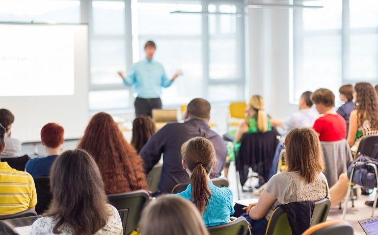 Fotografia colorida de uma sala de aula repleta de alunos e o professor. Os alunos, em primeiro plano, estão sentados de costas para a foto e de frente para o professor. O professor, em imagem desfocada, está de frente para os alunos com os braços flexionados e as mãos viradas para cima. Veste camisa azul e calça preta.