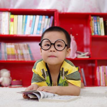 Fotografia colorida de um menino oriental deitado de bruços com os bracinhos flexionados sobre um livro aberto e sob um tapete branco. Ele olha para frente, usa óculos redondos e veste uma camiseta amarela com detalhes riscados em cinza e preto. Está em uma sala como uma estante vermelha repleta de livros atrás dele.