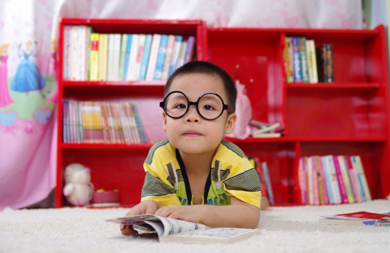 Fotografia colorida de um menino oriental deitado de bruços com os bracinhos flexionados sobre um livro aberto e sob um tapete branco. Ele olha para frente, usa óculos redondos e veste uma camiseta amarela com detalhes riscados em cinza e preto. Está em uma sala como uma estante vermelha repleta de livros atrás dele.