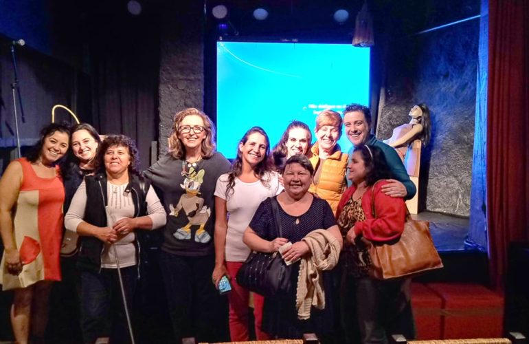 Fotografia colorida do grupo de atores com pessoas com deficiência e audiodescritores, logo após a peça. Da esquerda para direita: Andréia Paiva, Joselba Fonseca, Terezinha Aparecida, Livia Motta, Brisa Teixeira, Michelle Paim, Maria Lucia, Daniel Fernandes e Helen Mieko, na frente do palco do Basement Cultural.