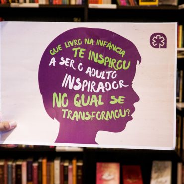 Banner de incentivo à leitura. Foto de uma estante de livro com um banner com fundo branco e a silhueta ao centro de um rosto de perfil na cor rocha escrito nas cores branca e verde "Que livro na infância te inspirou a ser o adulto inspirador no qual se transformou?"