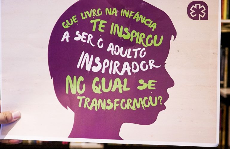 Banner de incentivo à leitura. Foto de uma estante de livro com um banner com fundo branco e a silhueta ao centro de um rosto de perfil na cor rocha escrito nas cores branca e verde "Que livro na infância te inspirou a ser o adulto inspirador no qual se transformou?"