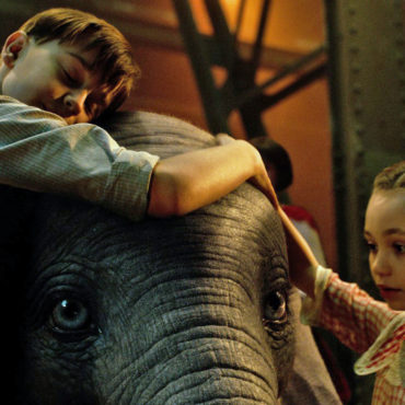 Dumbo ao centro é abraçado por menino e menina. Cena do filme Dumbo com Audiodescrição e Libras.