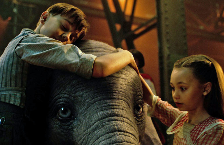 Dumbo ao centro é abraçado por menino e menina. Cena do filme Dumbo com Audiodescrição e Libras.