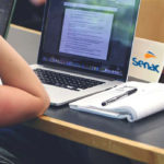 Curso de inclusão Senac: Foto de uma mesa de estudos com computador, caderno e caderno e caneta. Com uma pessoa estudando, em que aprece apenas o braço dela.