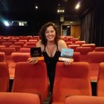 Audiodescritora Andréia Paiva na sala de cinema com estofados vermelho. Ela segura dois troféus do V Festival VerOuvindo