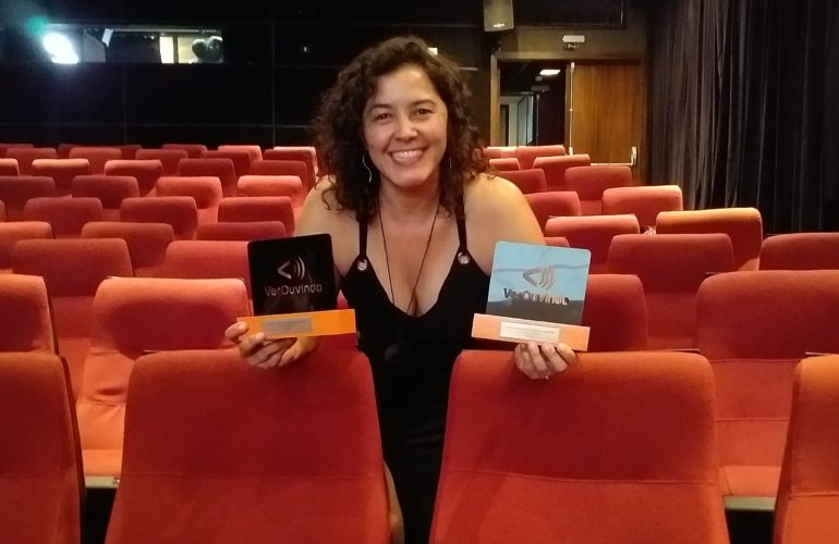 Audiodescritora Andréia Paiva na sala de cinema com estofados vermelho. Ela segura dois troféus do V Festival VerOuvindo