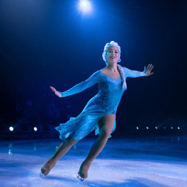 Audiodescrição: Elza patina sorridente de de braços abertos. Seu vestido é azul de mangas compridas. Narração da Audiodescriçãopor Raquel Carissimi