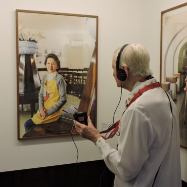 Idosa com fone de ouvido de audiodescrição está de costas e em frente a uma das fotos da exposição Observador, em que aparece uma mulher oriental posando para a foto.