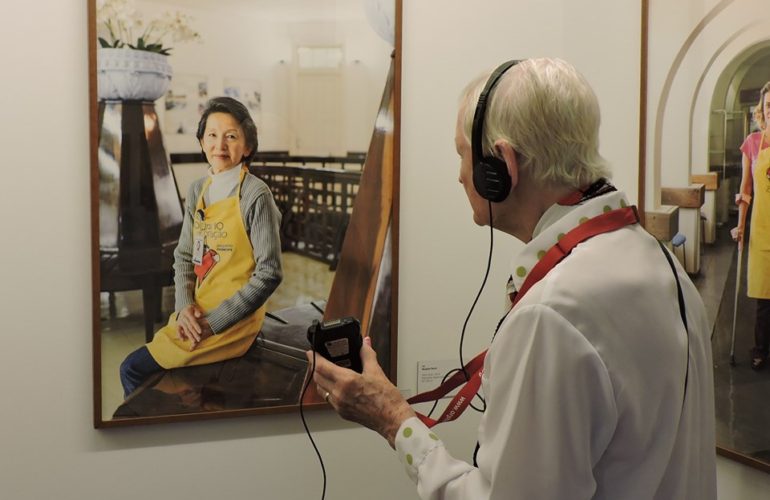 Idosa com fone de ouvido de audiodescrição está de costas e em frente a uma das fotos da exposição Observador, em que aparece uma mulher oriental posando para a foto.
