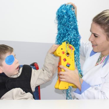 Adaptação escolar - Foto de menino com tampão no olho direito tateia objetos coloridos estimulado por uma professora. Ela sorri.