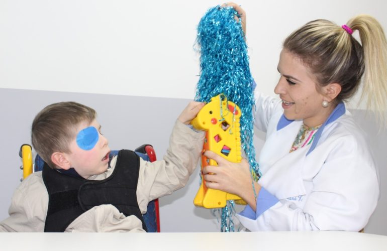 Adaptação escolar - Foto de menino com tampão no olho direito tateia objetos coloridos estimulado por uma professora. Ela sorri.