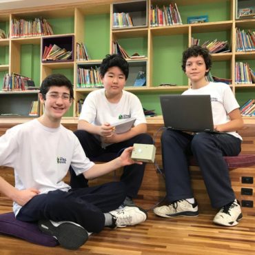 Três alunos sentados no chão de uma Biblioteca. Seguram notebook e um tablet.