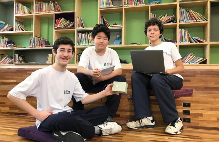 Três alunos sentados no chão de uma Biblioteca. Seguram notebook e um tablet.