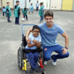Prática inclusiva Foto quadrada do menino na cadeira de rodas e o professor ao lado agachado e abraçado ao aluno.