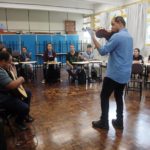 Foto do professor Luiz Amorim em uma sala de aula. Ele está de costas, toca violão para os alunos dispostos em semicírculo.