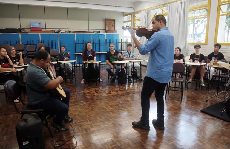 Foto do professor Luiz Amorim em uma sala de aula. Ele está de costas, toca violão para os alunos dispostos em semicírculo.